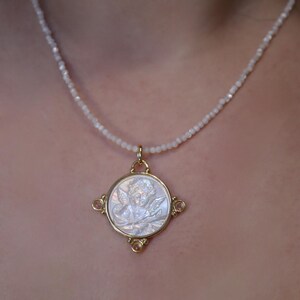 May include: A gold-toned pendant necklace featuring a detailed angel cameo. The pendant is set in a square frame with four small, pinkish-orange gemstones. The necklace has a delicate, beaded chain.