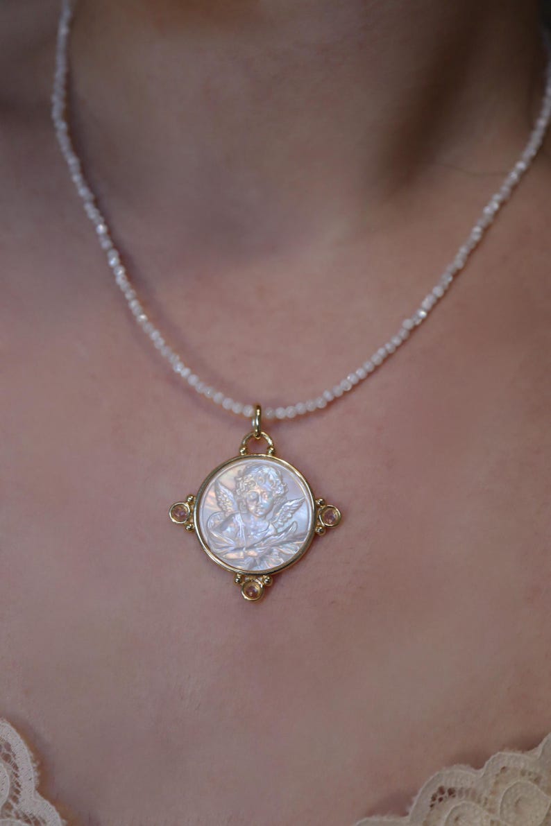 May include: A gold-toned pendant necklace featuring a detailed angel cameo. The pendant is set in a square frame with four small, pinkish-orange gemstones. The necklace has a delicate, beaded chain.