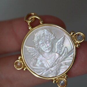 May include: A gold pendant featuring a carved angel design within a circular frame. The angel is depicted with wings and flowing garments, set against a pearlescent background. The pendant has three small, clear gemstones.