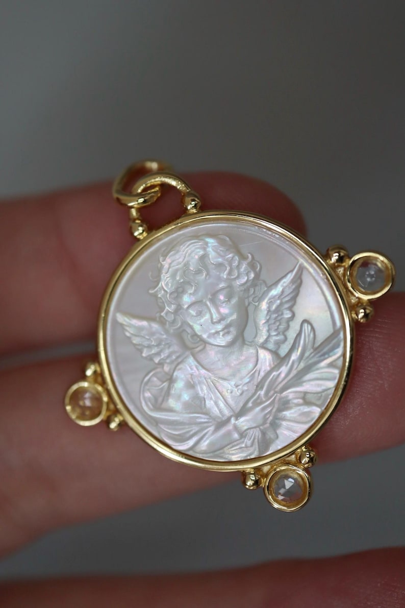 May include: A gold pendant featuring a carved angel design within a circular frame. The angel is depicted with wings and flowing garments, set against a pearlescent background. The pendant has three small, clear gemstones.
