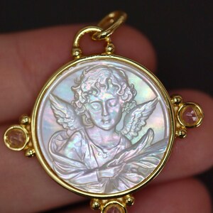 May include: A gold pendant with a carved mother-of-pearl cameo depicting an angel with wings. The pendant has a circular design with a gold frame and small pink gemstones at the corners. The angel is holding a branch.