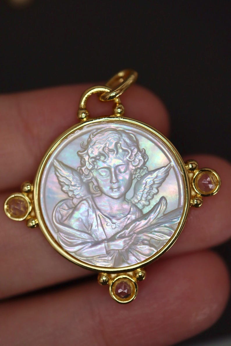 May include: A gold pendant with a carved mother-of-pearl cameo depicting an angel with wings. The pendant has a circular design with a gold frame and small pink gemstones at the corners. The angel is holding a branch.