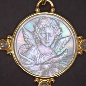 May include: A gold pendant featuring a detailed relief of an angel within a circular frame. The angel has wings and is holding an object. The pendant is accented with clear gemstones and a gold bail for hanging. The background is dark.