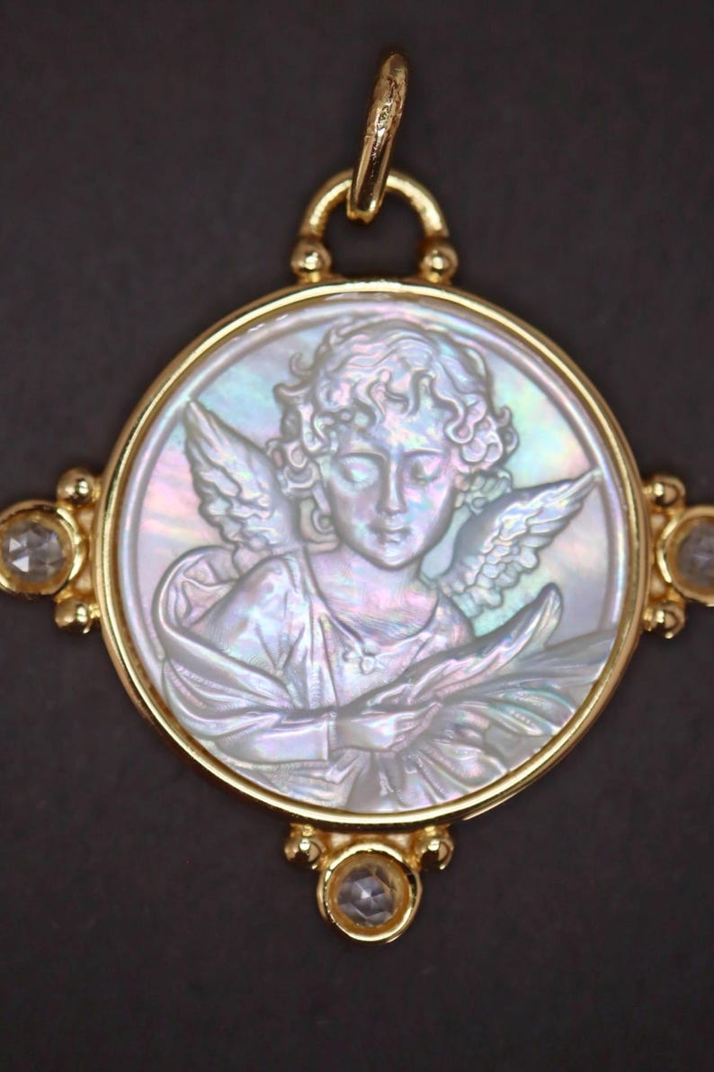 May include: A gold pendant featuring a detailed relief of an angel within a circular frame. The angel has wings and is holding an object. The pendant is accented with clear gemstones and a gold bail for hanging. The background is dark.