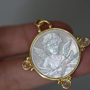 May include: Gold-tone pendant featuring a carved angel design within a circular frame. The angel is depicted with wings and a serene expression, set against a pearlescent background. The pendant has three small, clear gemstones.