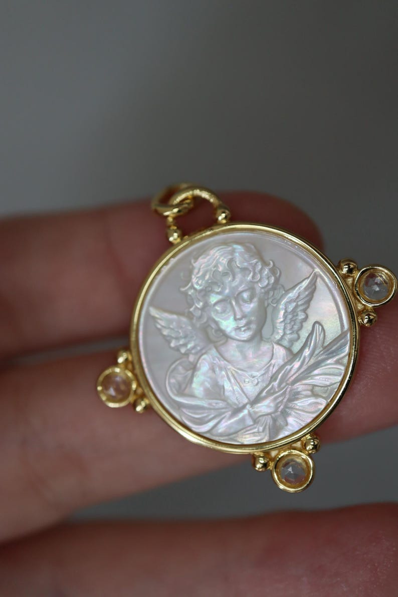May include: Gold-tone pendant featuring a carved angel design within a circular frame. The angel is depicted with wings and a serene expression, set against a pearlescent background. The pendant has three small, clear gemstones.