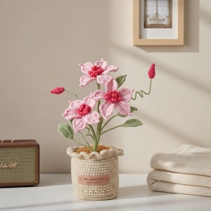 May include: A handmade crochet flower arrangement in a beige pot. The pink flowers have red centres and green leaves. The pot has a scalloped edge and a tag that reads "Handmade Limited edition."