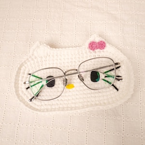 May include: Silver-framed eyeglasses rest on a white crocheted cat-shaped eyeglass holder. The holder features black, green, and yellow details, with a pink bow. The background is a white, textured surface.