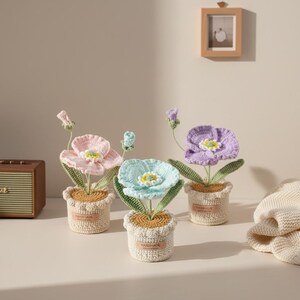 May include: Three handmade crochet flowers in small pots. The flowers are in shades of pink, blue, and purple, with yellow centers and green leaves. The pots are beige and have a handmade label. A small wooden frame hangs on the wall.