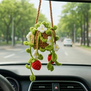 May include: A handmade crochet hanging car charm featuring a brown basket with green leaves, white flowers, and red strawberries. The charm is suspended by a brown rope, adding a touch of nature-inspired decor to the car's interior.