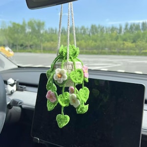 May include: A handmade crochet hanging planter with white, pink, and yellow flowers and green leaves. The planter is suspended by white cords and is hanging in a car. The crochet work is detailed and the colors are vibrant.