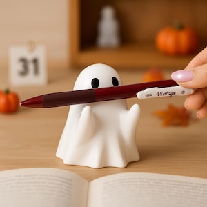 May include: A white ghost-shaped pen holder with black eyes and a maroon pen. The pen has the text "DS Vintage" on it. The pen holder is on a wooden surface with a calendar and small pumpkins in the background.