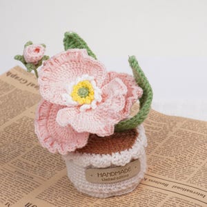 May include: A handmade crochet flower arrangement featuring a large pink flower with a yellow and white center, a small pink bud, and green leaves. The flower sits in a crocheted pot with a "HANDMADE Limited edition" tag.
