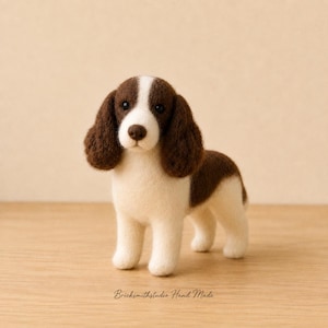 May include: A needle-felted dog figurine with brown and cream-coloured wool. The dog has long, floppy brown ears, a white blaze on its face, and dark eyes. The text "Bricksmithstudio Hand Made" is printed in small, dark letters at the bottom.