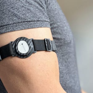 Puede incluir: Un dispositivo médico negro y gris está sujeto a un brazo. El dispositivo tiene una cara circular con la palabra "Dexcom" impresa. La correa es negra y ajustable. El brazo está cubierto por una camiseta gris.