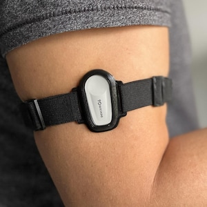 Op de afbeelding: Een zwart-witte Dexcom G6 continue glucose monitoring (CGM) sensor is met een zwarte band aan de arm bevestigd. Dit medische hulpmiddel wordt gebruikt om de glucosewaarden te controleren.