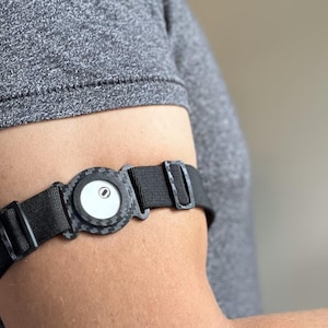 Könnte beinhalten: Ein schwarzer Fitnesstracker mit rundem Zifferblatt und schwarzem Armband wird am Arm getragen. Der Tracker hat eine weiße Mitte mit einem kleinen schwarzen Punkt. Das Armband besteht aus gewebtem Material und hat eine Schnalle.