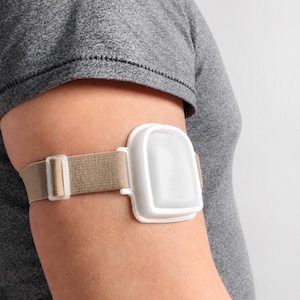 Op de afbeelding: Een witte, rechthoekige behuizing voor een medisch apparaat met afgeronde hoeken, bevestigd aan de bovenarm door een beige elastische band met een witte plastic verstelgesp. Het apparaat heeft een gladde, witte afdekking en een platte basis.