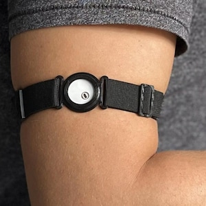 Op de afbeelding: Een zwarte armband met een cirkelvormig apparaat. Het apparaat heeft een witte voorkant met een kleine zwarte stip. De band wordt vastgemaakt met een zwarte gesp. Het item wordt op de bovenarm gedragen.