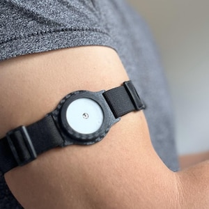 May include: A black wearable device with a circular face and a white centre is secured to an arm with a black strap. The device is worn on the upper arm, and the strap is fastened with a buckle.