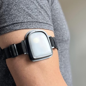 Può includere: Un dispositivo indossabile nero e grigio è legato a un braccio. Il dispositivo ha una forma rettangolare con bordi arrotondati e uno schermo bianco brillante. Il cinturino è nero e regolabile. La persona indossa una maglietta grigia.