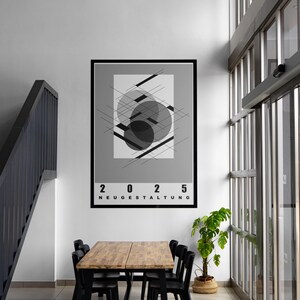 May include: A framed poster with a geometric design in black and gray, featuring overlapping shapes and lines. The text "2025 NEUGESTALTUNG" is displayed at the bottom. The poster hangs above a wooden table and black chairs.