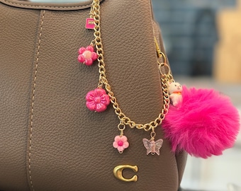 Puede incluir: Bolso de mano de cuero color topo con una cadena dorada y abalorios de flores rosas. Un pompón rosa y un logotipo "C" dorado añaden elementos decorativos. El bolso tiene un asa curva y un diseño cosido.