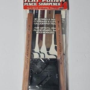 Op de afbeelding: Een General's Flat Point puntenslijper in een doorzichtige plastic verpakking met een rode kop. De set bevat twee houten schetspotloden en een zwarte plastic puntenslijper met metalen mesjes. De verpakking bevat de tekst NEW, FLAT POINT PENCIL SHARPENER en FOR ARTISTS, CRAFTSPEOPLE, CALLIGRAPHERS, AND WOODWORKERS.