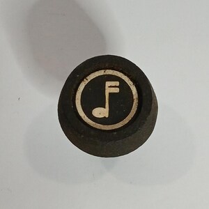 May include: A circular black knob featuring a centred inlay with a light-coloured musical note symbol. The knob has a textured, dark surface and a distinct circular border surrounding the graphic icon, presenting a simple and functional design.