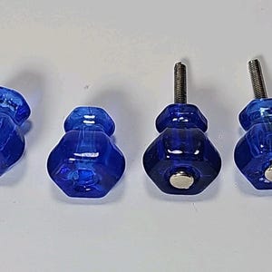 以下が含まれることがあります： ファセット加工された六角形のコバルトブルーのガラス製キャビネットノブ4個。2個のノブには背面に銀色の金属製ネジが突き出ており、残りの2個には前面に平らな銀色の金属製ベースが付いています。