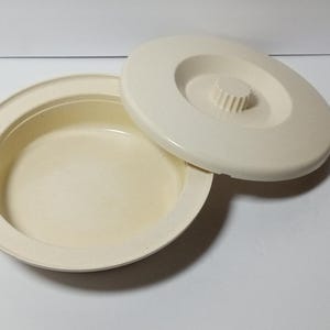 May include: Off-white, round container with a matching lid. The container has a shallow, bowl-like shape with a raised rim. The lid features a central, raised knob. The items are made of a solid, opaque material.