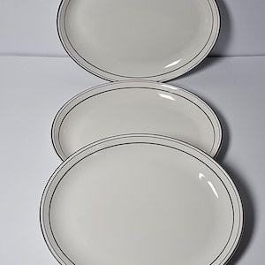 May include: A set of three round, off-white ceramic plates stacked vertically. Each plate features a thin, dark black circular band running along the outer rim, providing a simple contrast against the smooth, light-colored surface of the dinnerware.