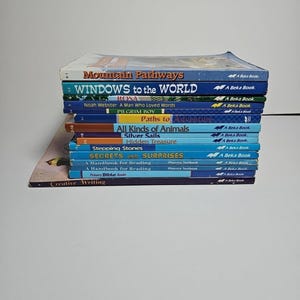 May include: A stack of colorful children's books with titles like "Mountain Pathways," "Windows to the World," and "Secrets and Surprises." The books are various sizes and colors, with text visible on the spines.