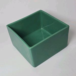 May include: A square-shaped ceramic bowl with a smooth, glossy finish in a solid sage green colour. The container features straight, vertical walls and a flat base, presenting an open, hollow interior space with clean, sharp edges throughout.