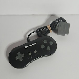 May include: A black Hyperkin video game controller featuring a gray directional pad, four gray action buttons, and two smaller gray buttons labeled Select and Start. The controller includes a black cord with a gray connector plug attached.