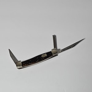 May include: A folding pocket knife with three distinct metal blades extended. The handle features a dark brown, textured, jigged bone-style grip with a small, silver-colored metal shield inlay embedded in the center of the handle scales.