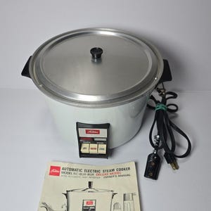 Può includere: Una vaporiera elettrica automatica Toshiba bianca con coperchio argentato, manici laterali neri e un pannello di controllo con pulsanti Warm e Cook. Un cavo di alimentazione nero e un manuale intitolato Automatic Electric Steam Cooker.