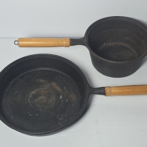 May include: Two black cast iron cooking pots: a frying pan and a small saucepan. Both have light-coloured wooden handles. The frying pan is round with a single handle and a small loop handle. The saucepan has a single handle.