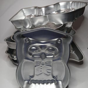 May include: A stack of silver-colored metal baking pans. The front pan is shaped like an owl wearing a graduation cap and bow tie. Other pans have various shapes, including a rectangular one with scalloped edges. The pans are stacked on a white surface.