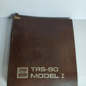 Może przedstawiać: Brązowa książka w skórzanej oprawie z napisem "TRS-80 MODEL I" i "Radio Shack TRS-80 Software Library" wydrukowanym na okładce. Książka ma zużyty wygląd.