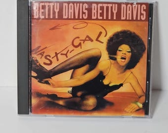 May include: A CD case featuring the album cover for Betty Davis's "Nasty Gal." The cover art shows a woman in a black dress and fishnet stockings, with the album title and artist's name in a brown font.