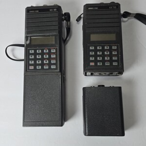 May include: Two black Bendix/King KX 99 handheld transceivers and one separate black battery pack. Each radio features a digital display, a numeric keypad with function buttons, and a black wrist strap. The devices are rectangular in shape.