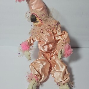 Puede incluir: Muñeca vintage vestida con un disfraz de payaso de satén. La muñeca lleva un mono de satén rosa, un sombrero a juego con un pompón rosa y un cuello con volantes. Las manos y los pies de la muñeca son blancos con detalles rosas.