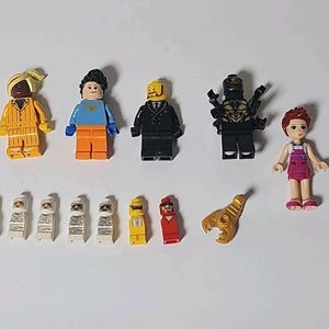 Pode incluir: Uma coleção de figuras de blocos de montar de plástico, incluindo cinco minifiguras padrão com várias roupas, uma figura mais alta com um vestido roxo, cinco pequenas figuras brancas tipo múmia, duas pequenas figuras coloridas e um acessório de garra dourada.