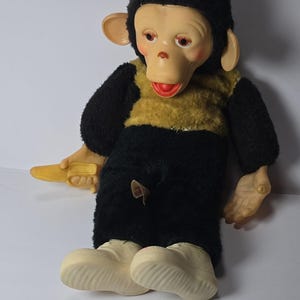 May include: A plush monkey doll with black fur on the head and limbs, a tan chest, and a light-coloured face with large ears. The doll features moulded plastic hands and feet, a yellow banana accessory in one hand, and a small tag on the torso.