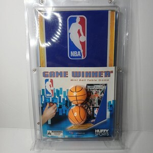 May include: A Huffy Sports NBA Game Winner mini ball table game in clear plastic packaging. The blue and white box features the NBA logo, the text Game Winner, and two orange basketballs. The packaging includes a warning label at the bottom.