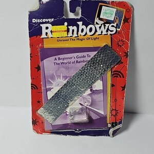 May include: A rectangular diffraction grating tool with a textured, grid-like surface, packaged on a purple and red cardboard backing. The packaging features the text Discover Rainbows Unravel The Magic of Light and A Beginner's Guide To The World of Rainbows.
