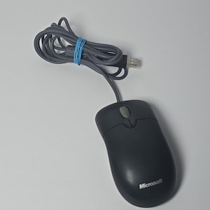 May include: A black Microsoft computer mouse with a gray scroll wheel and a long gray USB cable secured with a blue rubber band. The device features a smooth, ergonomic shape with the Microsoft logo printed in white on the lower right side.