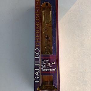 May include: A Galileo thermometer in a box. The thermometer is 17 inches tall and made of handcrafted blown glass with 24K gold plated medallions. The box is dark blue and burgundy with the text "GALILEO THERMOMETER".