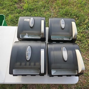 May include: Four black paper towel dispensers with oval-shaped viewing windows and white and silver accents. The dispensers are arranged on a white surface against a backdrop of green grass.